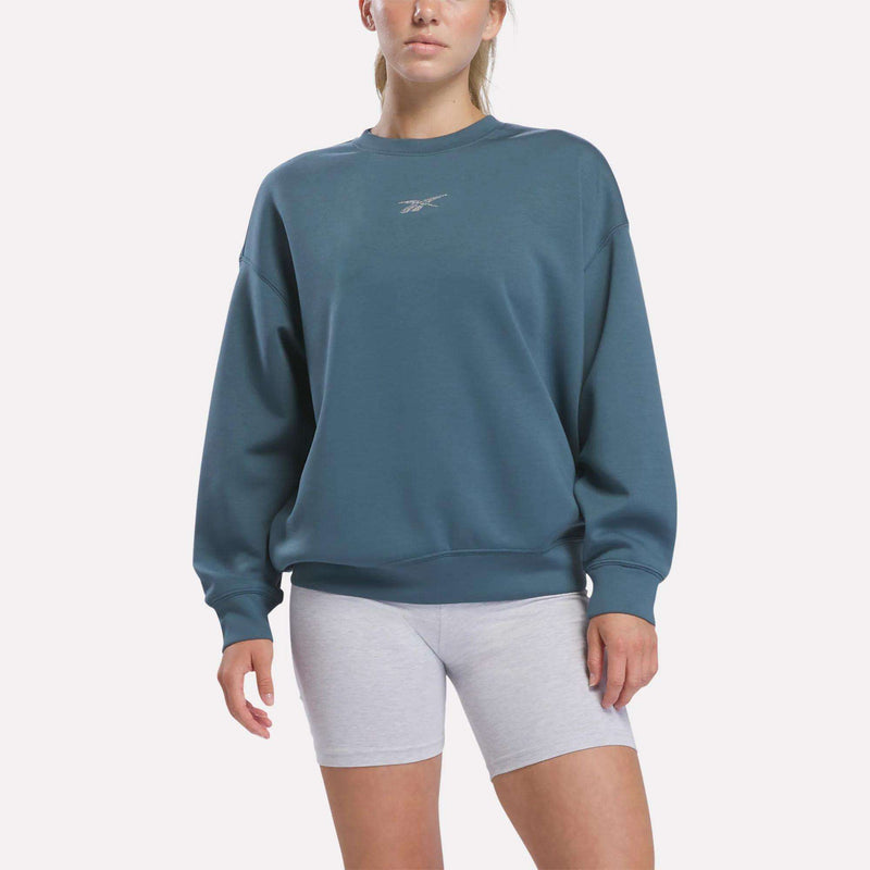 A person stands against a plain background wearing the Reebok Women’s Relaxed Crewneck Sweatshirt in Midnight Tide, which features a relaxed silhouette and a small logo on the chest, paired with light gray shorts.