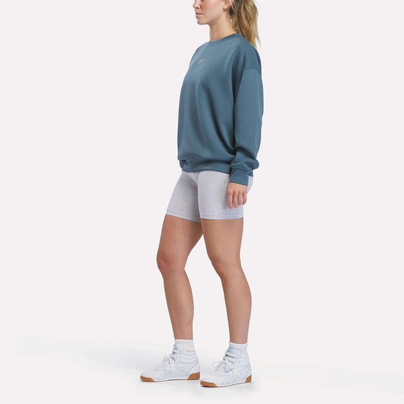 A person stands sideways against a plain white background, wearing a teal Reebok Women’s Relaxed Crewneck Sweatshirt with a roomy silhouette, paired with light gray bike shorts, white socks, and white sneakers.