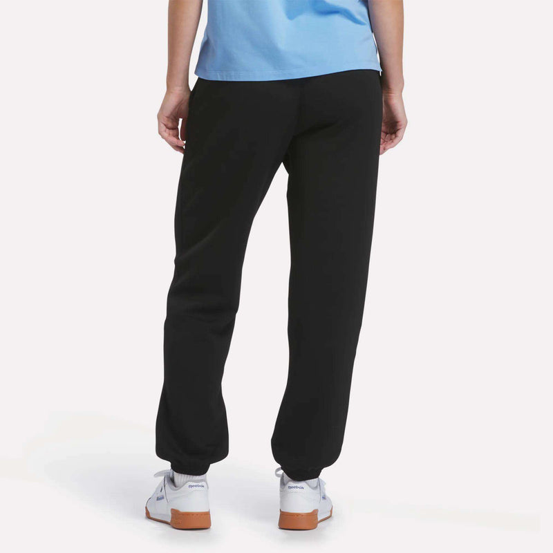 A person stands facing away against a plain white background, wearing a light blue shirt, Reebok Women’s Cinched Joggers in soft black, and white sneakers with brown soles.
