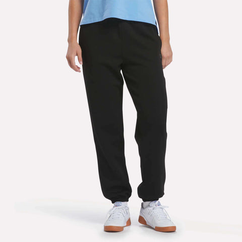 A person wearing Reebok Women’s Cinched Joggers in black, a light blue shirt, and white sneakers stands with arms relaxed at their sides against a plain white background.