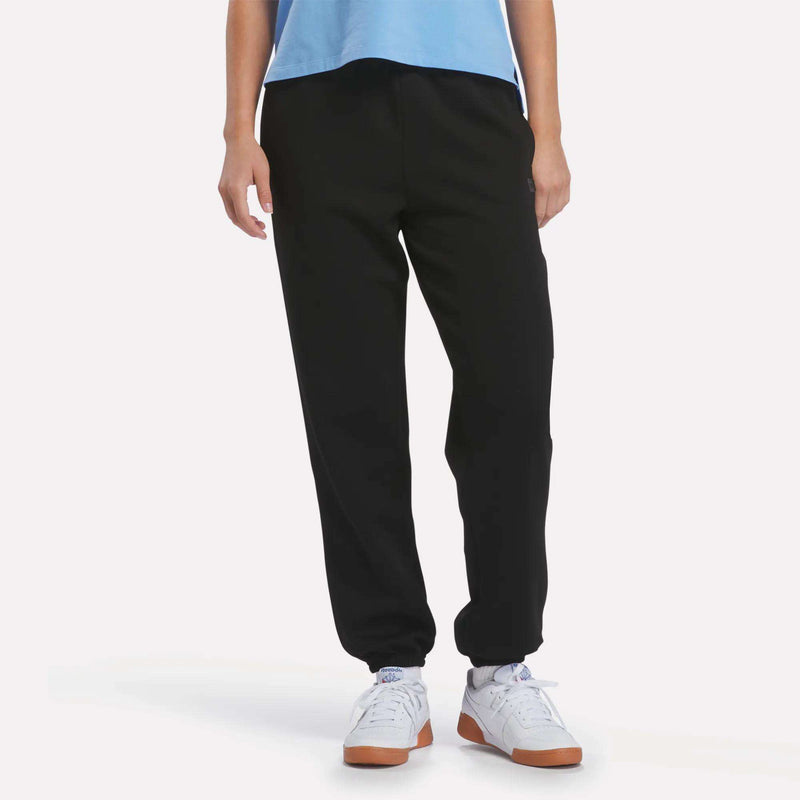 A person wearing Reebok Women’s Cinched Joggers in black, a light blue shirt, and white sneakers stands with arms relaxed at their sides against a plain white background.