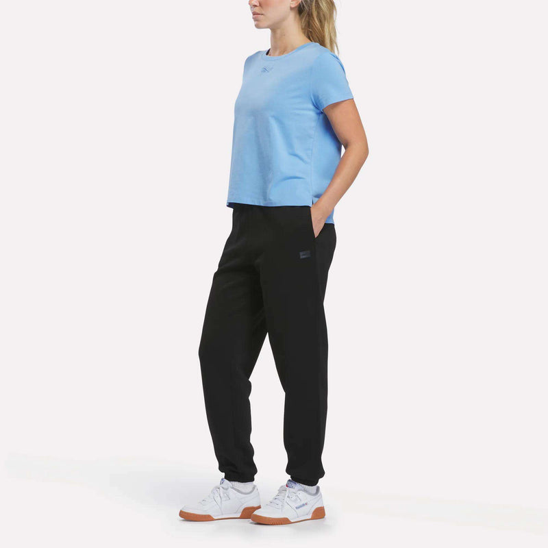 A woman stands against a white background, wearing a light blue t-shirt, Reebok Women’s Cinched Joggers in black, and white sneakers with gum soles. Her hands are in her pockets and she looks off to the side.