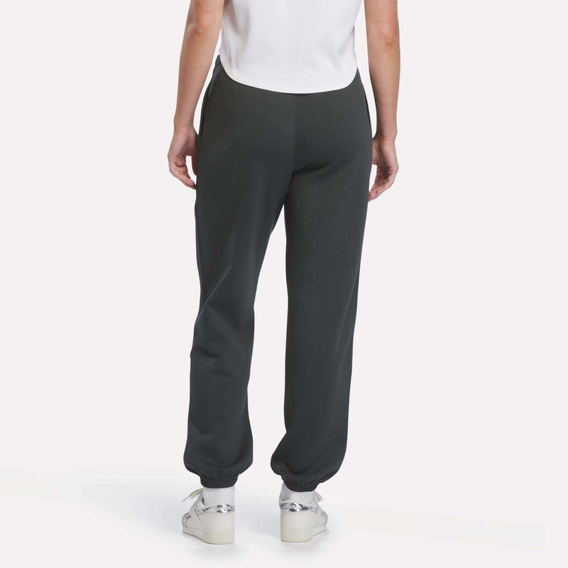 A person is shown from the back, standing against a plain white background, wearing a white shirt, Reebok Women’s Cinched Joggers in dark gray, and white sneakers.