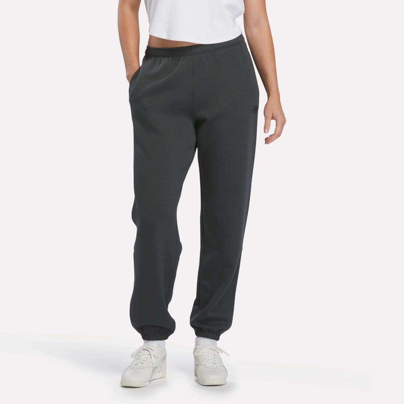A person wearing a white t-shirt, Reebok Women’s Cinched Joggers in Grey 6, and white sneakers stands with hands in pockets against a plain light background.
