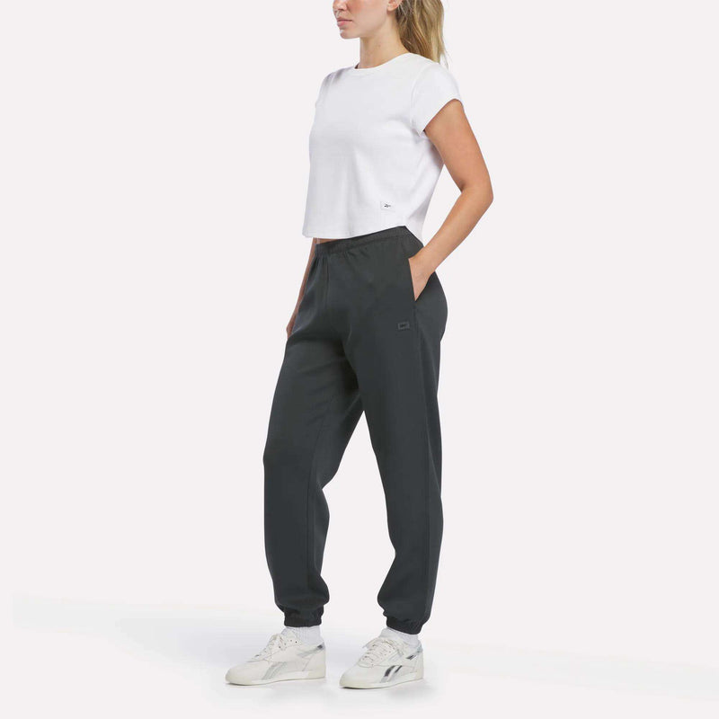 A person wearing a white cropped t-shirt, Reebok Women’s Cinched Joggers, and white sneakers stands with hands in pockets against a plain white background.