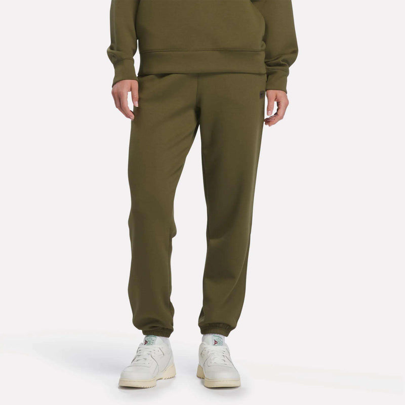 A person wearing Reebok Women’s Cinched Joggers in Olive Stone, a matching sweatshirt, and white sneakers stands against a plain white background.