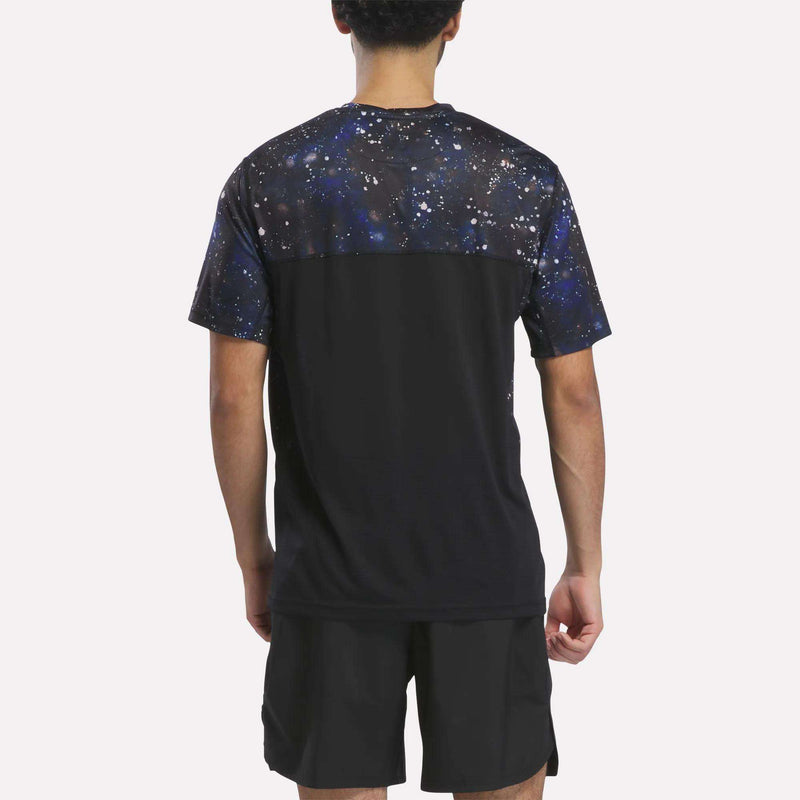 A man is seen from behind, wearing the Reebok Men’s Running Short Sleeve Tee in black, featuring a striking galaxy print across the upper back and shoulders, paired with black shorts. Against a plain white background, this versatile Reebok shirt blends style and performance for both everyday activities and workouts.