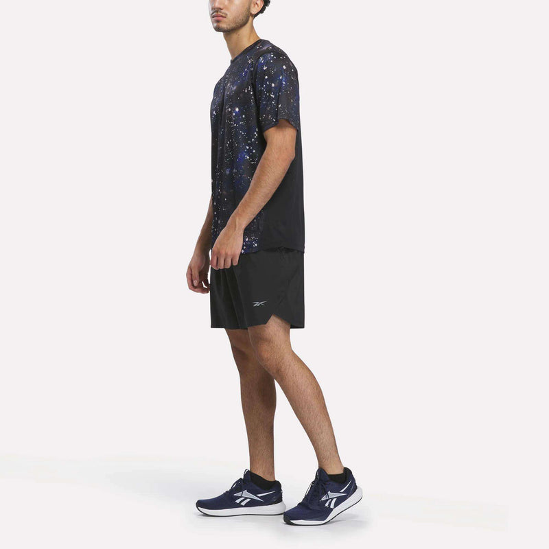 A person stands sideways against a plain white background wearing the Reebok Men’s Running Short Sleeve Tee—a black T-shirt featuring a blue and white galaxy pattern—paired with black shorts and navy blue Reebok sneakers.