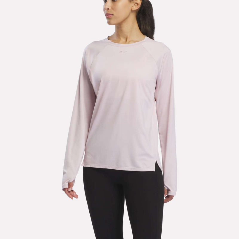 A woman in a Reebok Women’s Long Sleeve Running Top, Elemental Pink Heather, and black leggings stands facing slightly left against a plain white background.