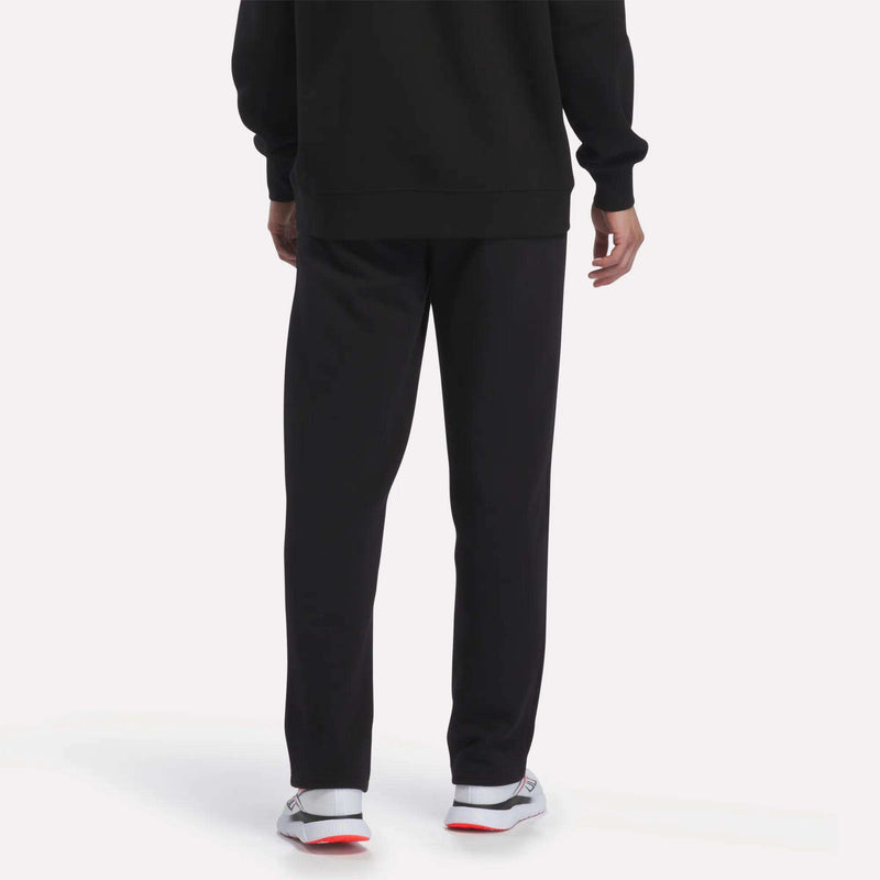 A person is standing and facing away from the camera against a plain background, wearing a black sweatshirt, white sneakers with red soles, and Reebok Men’s Open Hem Pants in black.