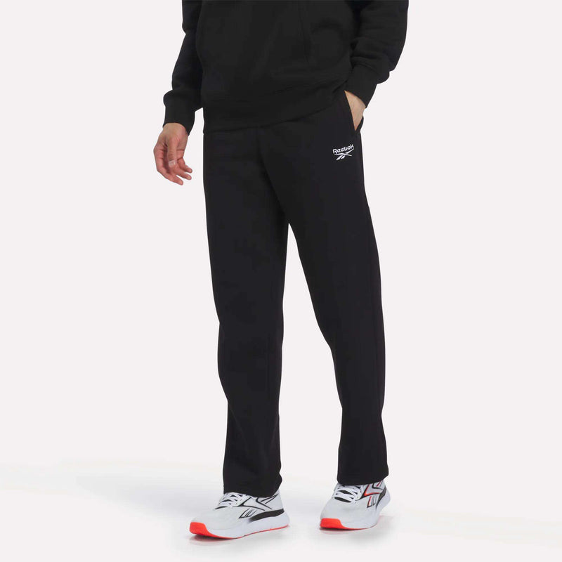 A person stands with one hand in their pocket against a plain background, wearing Reebok Men’s Open Hem Pants in black and a black sweatshirt, paired with white athletic shoes featuring red soles.