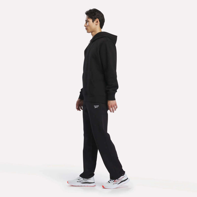 A person stands in profile wearing a black hoodie, white sneakers with red accents, and men’s Open Hem Pants from Reebok, against a plain white background.