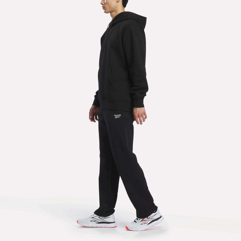 A man in a black hoodie and relaxed Reebok Men’s Open Hem Pants with a logo on the left thigh, wearing white athletic sneakers.