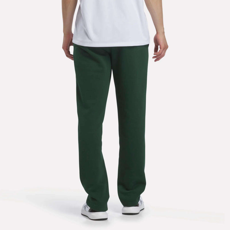 A person stands with their back to the camera against a plain white background, showcasing a relaxed look in a white T-shirt and Reebok Men’s Open Hem Pants in dark green, paired with white sneakers.