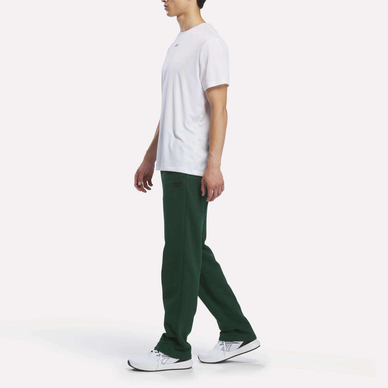 A person stands in profile against a white background, effortlessly showcasing a relaxed look while wearing a plain white t-shirt, Men’s Open Hem Pants from Reebok in dark green, and white sneakers with black accents.