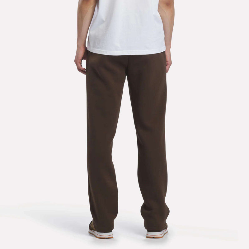 A person is standing and facing away from the camera, wearing a white T-shirt and Reebok Men’s Open Hem Pants in brown. Only their lower back, legs, and arms are visible against a plain, light-colored background.