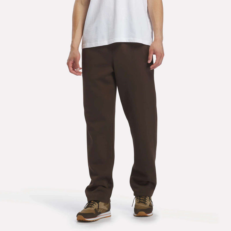 A person wearing a white T-shirt, Reebok Men’s Open Hem Pants in Espresso, and brown sneakers stands against a plain light background. The photo shows them from shoulders to feet.
