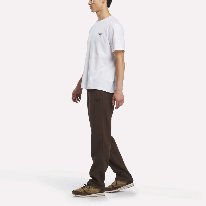 A person stands sideways against a plain white background, wearing a white T-shirt paired with Reebok Men’s Open Hem Pants in brown and brown sneakers, creating a casual and relaxed look.