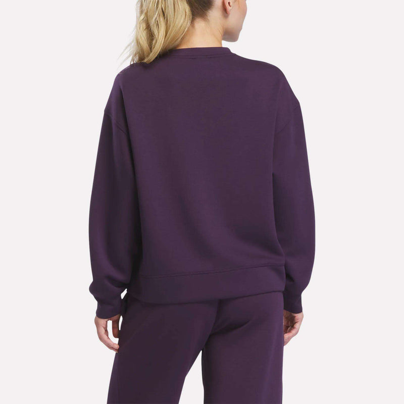 A woman with blonde hair in a ponytail is shown from the back, wearing a loose-fitting Reebok Women’s Crewneck Sweatshirt and matching purple pants against a plain light background.