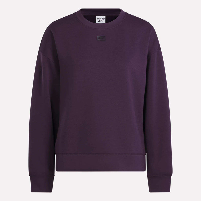 A women’s crewneck sweatshirt by Reebok, featuring long sleeves and a dark purple color. Designed with soft doubleknit fabric for comfort, it showcases a small black Reebok logo patch centered near the collar, blending everyday style and ease.