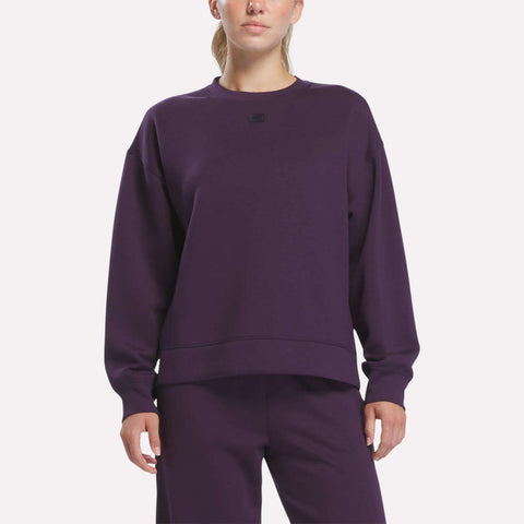 A woman stands against a plain white background wearing matching Reebok apparel in Phantom Purple. She is dressed in the Women’s Crewneck Sweatshirt by Reebok, which features long sleeves, a relaxed fit, and is crafted from comfortable doubleknit fabric.