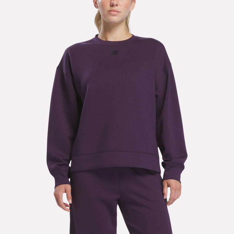 A woman stands against a plain white background wearing matching Reebok apparel in Phantom Purple. She is dressed in the Women’s Crewneck Sweatshirt by Reebok, which features long sleeves, a relaxed fit, and is crafted from comfortable doubleknit fabric.