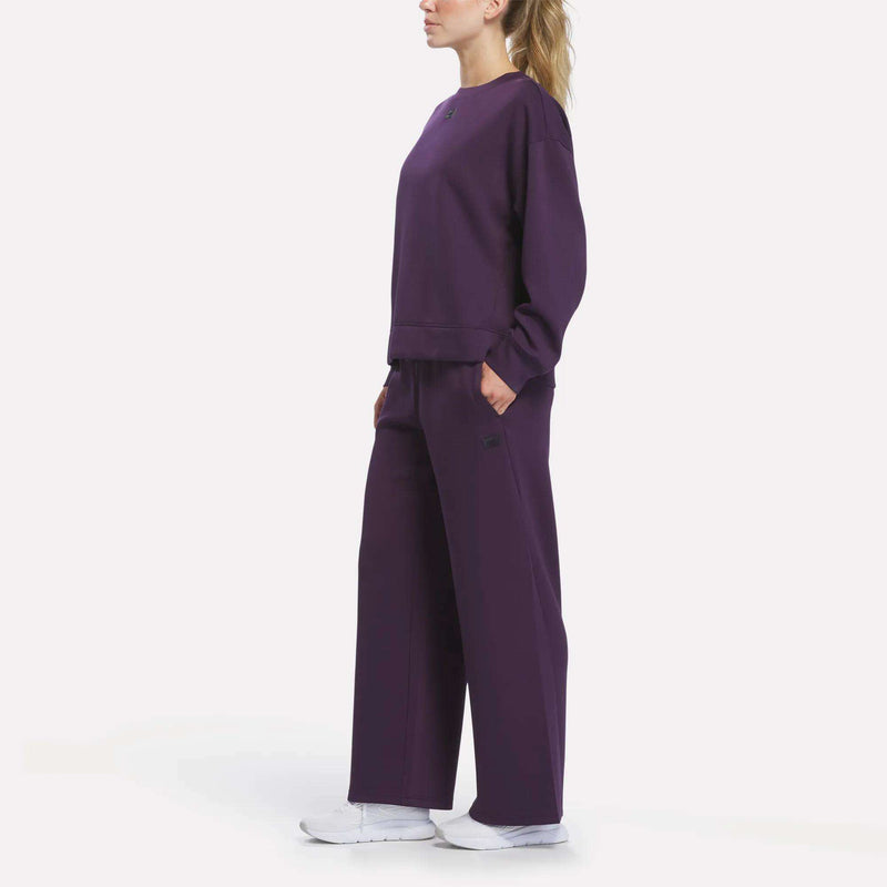 A woman stands in profile against a plain white background, wearing the Reebok Women’s Crewneck Sweatshirt and matching wide-leg pants in dark purple with her hands in her pockets, paired with white sneakers.