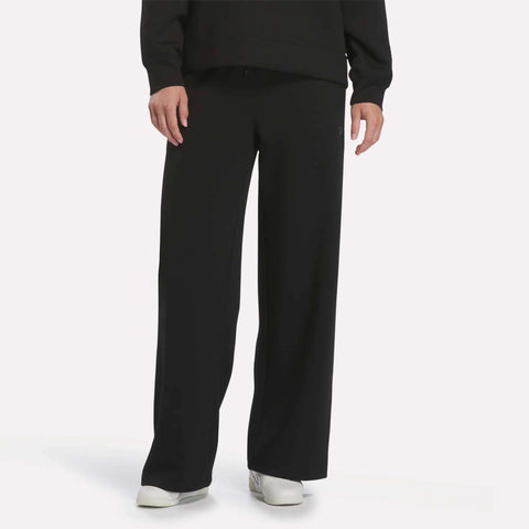 A person wearing Reebok Women’s Mid-Rise Wide-Leg Pants in black, white shoes, and a long-sleeve black top stands against a plain white background.