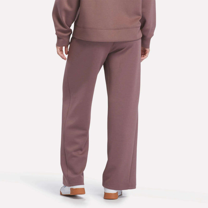 A person stands facing away from the camera, wearing Reebok Women’s Mid-Rise Wide-Leg Pants in mauve with a comfortable pull-on waistband and a matching sweatshirt, paired with white socks and brown-soled shoes against a plain light background.