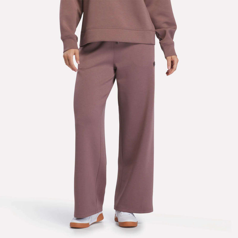 A person is wearing Reebok Women’s Mid-Rise Wide-Leg Pants in Light Cocoa, featuring a comfortable pull-on waistband, paired with a matching sweatshirt and white sneakers with tan soles, standing against a plain white background.