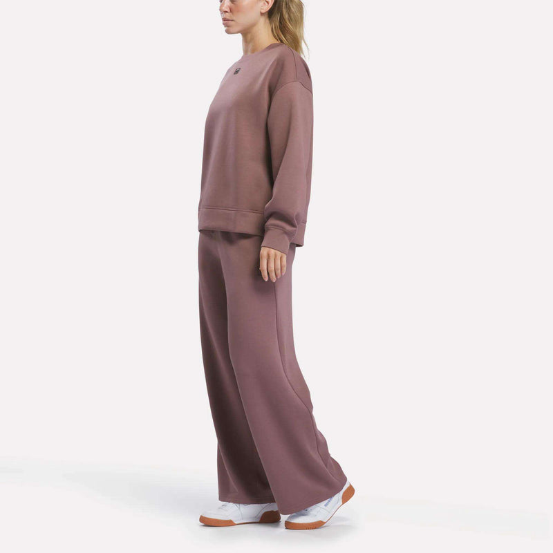 A person stands wearing mauve Reebok Women’s Mid-Rise Wide-Leg Pants with a pull-on waistband, paired with a loose matching sweatshirt and white sneakers. The background is plain white.