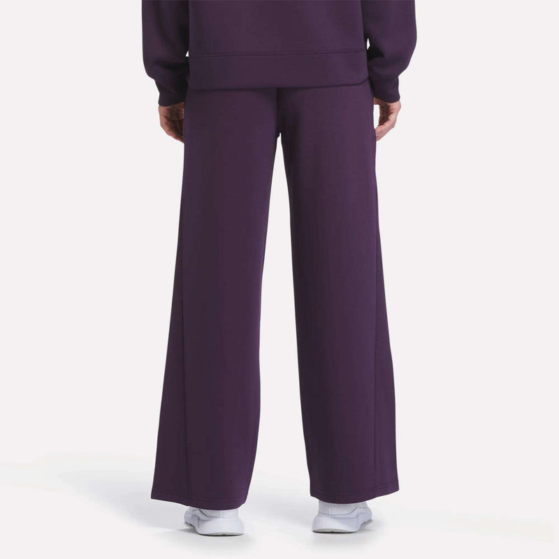 A woman is shown from the back wearing Reebok Women’s Mid-Rise Wide-Leg Pants in purple, styled with a matching purple sweatshirt and white sneakers, standing against a plain white background.