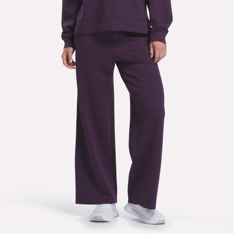 A person is shown from the waist down wearing Reebok Women’s Mid-Rise Wide-Leg Pants in Phantom Purple, paired with a matching purple sweatshirt and white sneakers, standing against a plain white background.