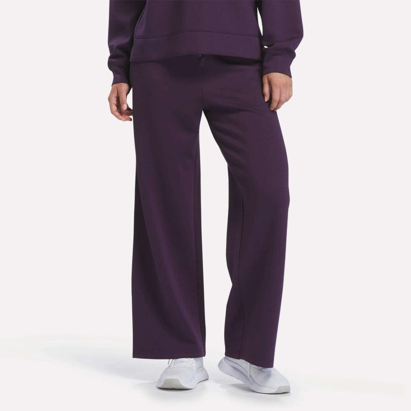 A person is shown from the waist down wearing Reebok Women’s Mid-Rise Wide-Leg Pants in Phantom Purple, paired with a matching purple sweatshirt and white sneakers, standing against a plain white background.