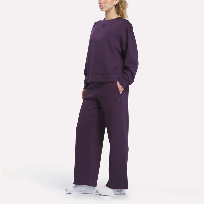 A woman stands with her hands in her pockets, wearing Reebok Women’s Mid-Rise Wide-Leg Pants in dark purple, paired with a matching sweatshirt and white sneakers. Her blonde hair is tied back, and she is posed against a plain white background.