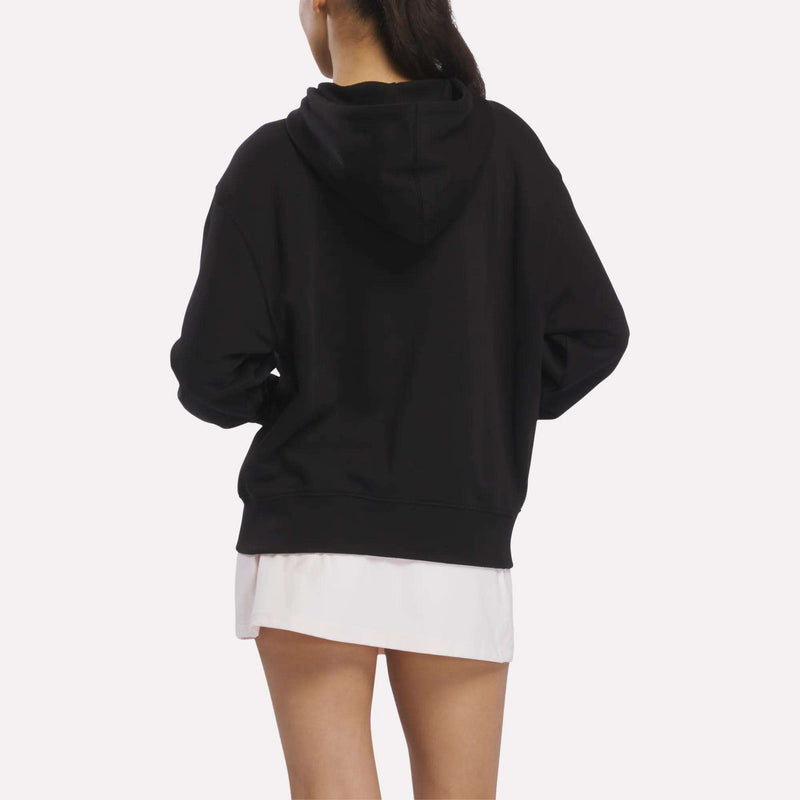 A person faces away, wearing a Reebok Women’s Oversized Full-Zip Hoodie over a white shirt against a plain, light-colored background.