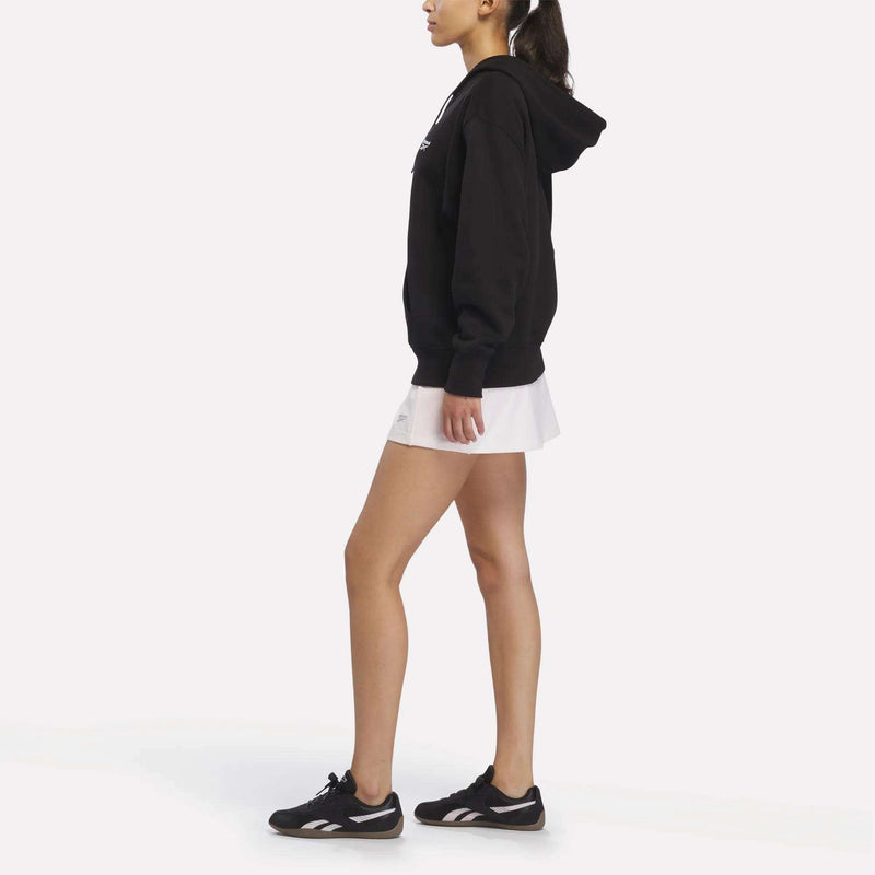 A person in profile wears a Women’s Oversized Full-Zip Hoodie by Reebok, paired with a white skirt and black Reebok sneakers, set against a plain white background.