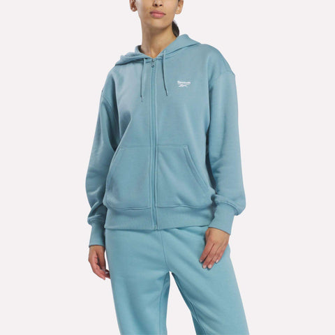 Woman in a Reebok Women’s Oversized Full-Zip Hoodie, Hazy Teal, standing with one hand in pocket, facing forward against a plain white background.