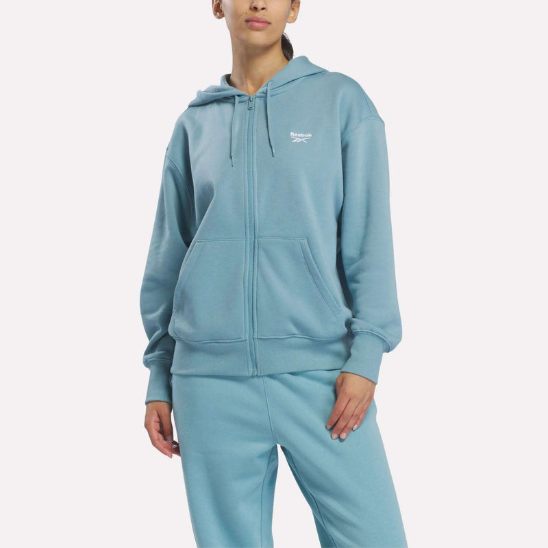 Woman in a Reebok Women’s Oversized Full-Zip Hoodie, Hazy Teal, standing with one hand in pocket, facing forward against a plain white background.