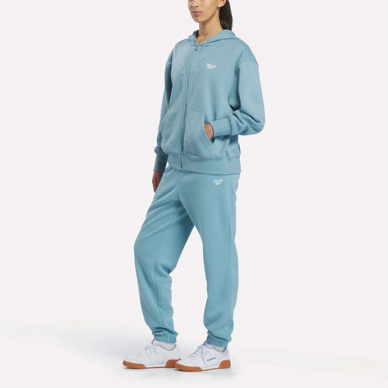 A person wears the Reebok Women’s Oversized Full-Zip Hoodie in light blue with matching sweatpants and white sneakers, standing against a plain white background.