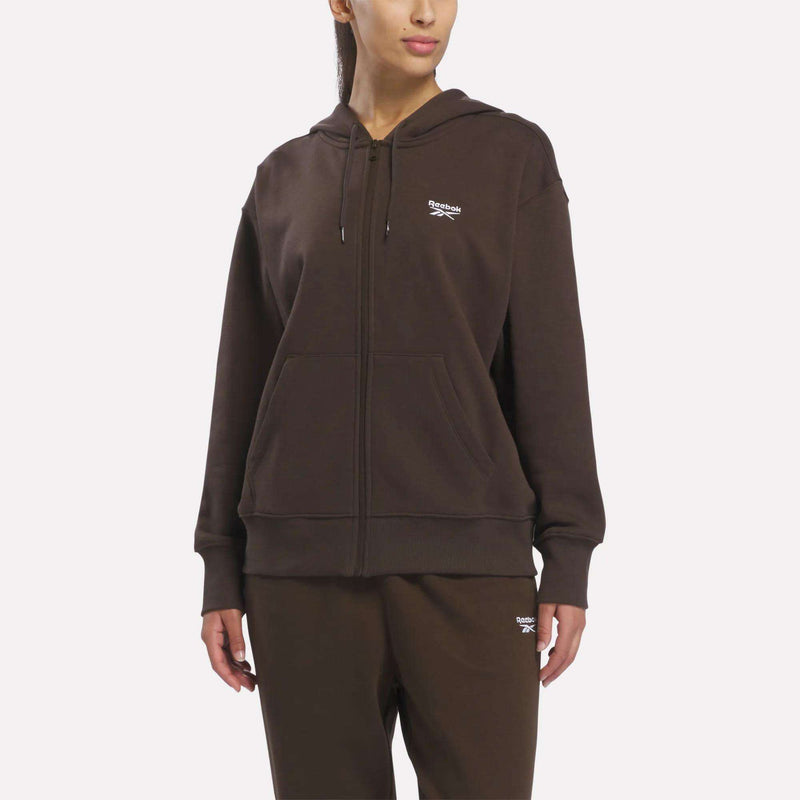 A person wears a Reebok Women’s Oversized Full-Zip Hoodie in Vector Espresso, paired with matching dark brown sweatpants featuring small white logos, styled against a plain light background.