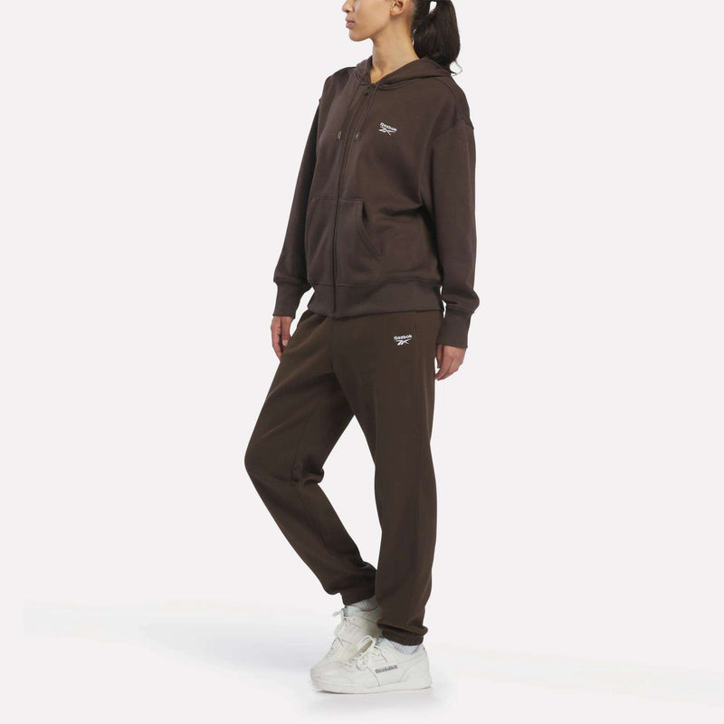 A person wears the Women’s Oversized Full-Zip Hoodie by Reebok in brown with small white logos, paired with matching sweatpants and white sneakers, against a plain white background.