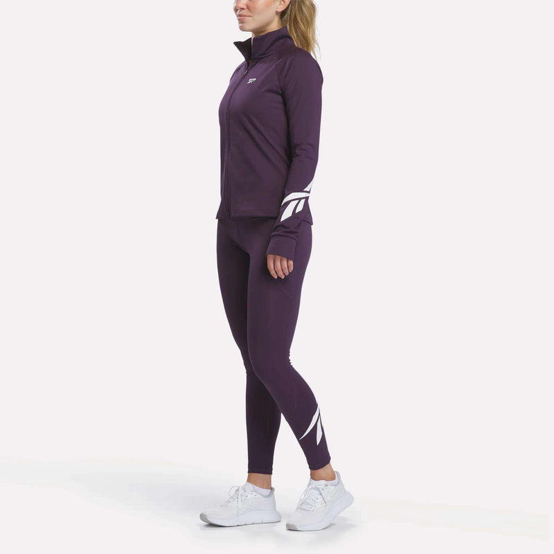 A person wearing the Reebok Women’s Motion Core Full-Zip in a purple athletic tracksuit with RBK-DRY technology—featuring white patterns on the sleeves and legs—and paired with white sneakers, stands against a plain white background.