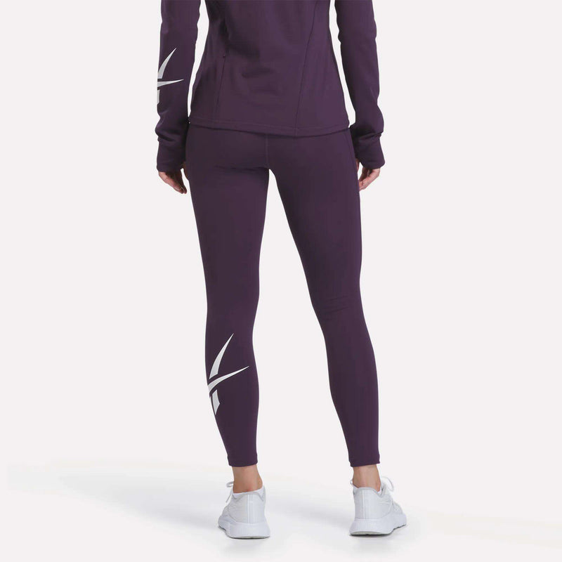A person stands with their back to the camera wearing Reebok Women’s Logo High-Rise Leggings featuring white graphic accents on the calves, paired with a purple athletic jacket with white detailing on the sleeves and white shoes.