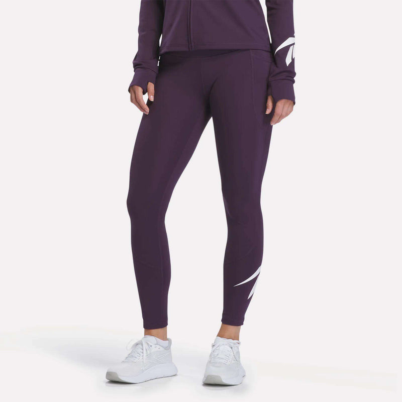 A person wearing Reebok Women’s Logo High-Rise Leggings in Phantom Purple, paired with a matching purple jacket and white sneakers, stands against a plain white background.