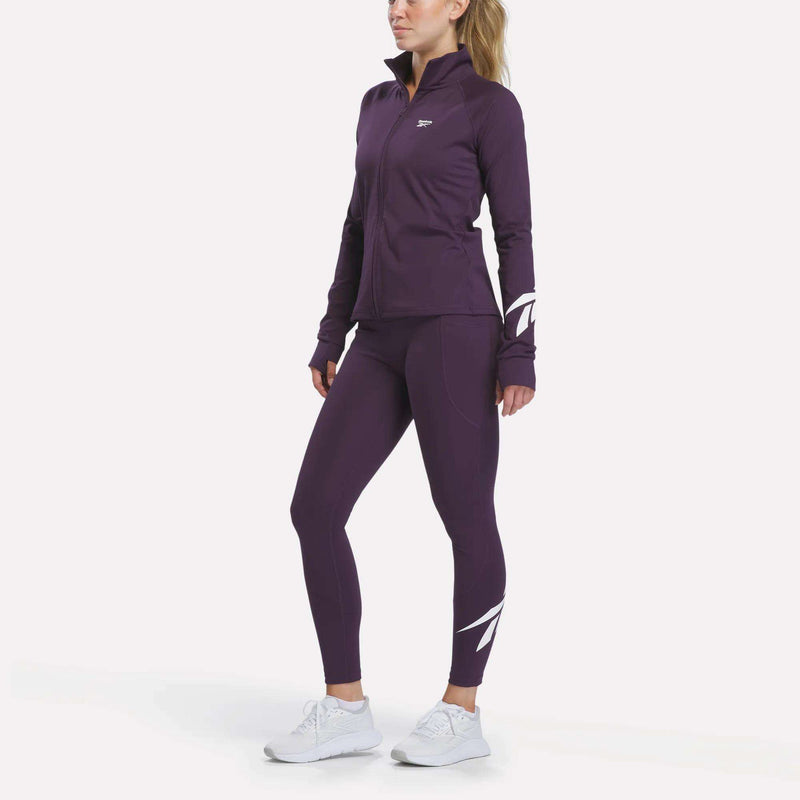 A woman stands against a plain white background wearing Reebok Women’s Logo High-Rise Leggings in purple, paired with a matching zip-up jacket, white athletic shoes, and her hair styled in a ponytail.