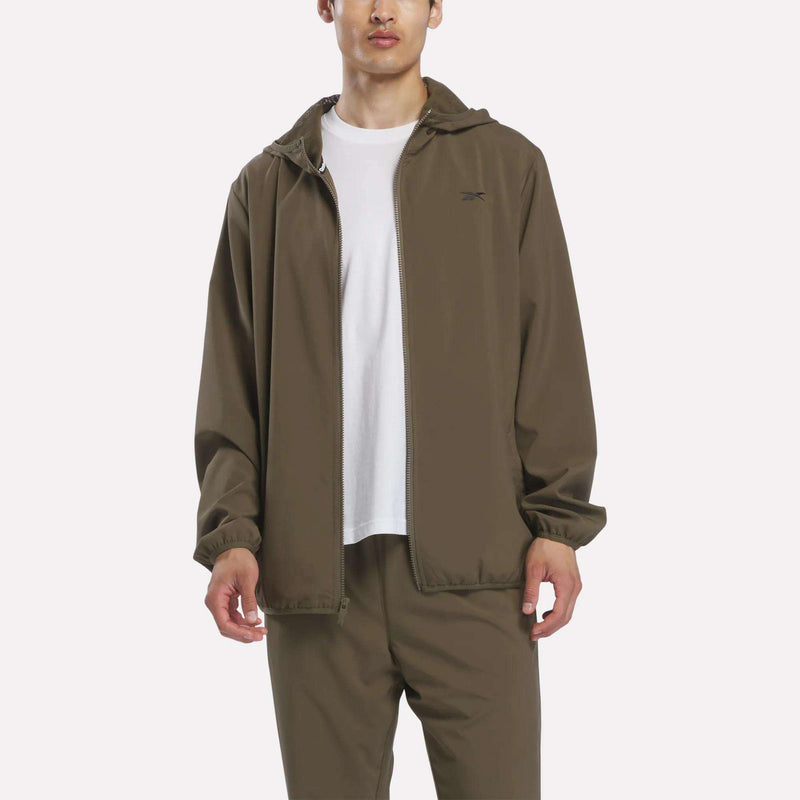 A man wearing a Reebok Men’s Woven Jacket in army green over a white t-shirt stands against a plain, light background.