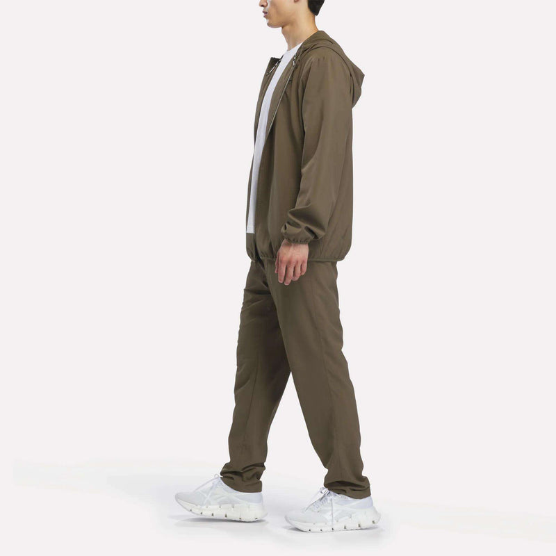 A man dressed in a brown Reebok Men’s Woven Jacket with a hood, paired with a white T-shirt and white sneakers, walks to the left against a plain white background, modeling stylish and athletic men’s training gear.