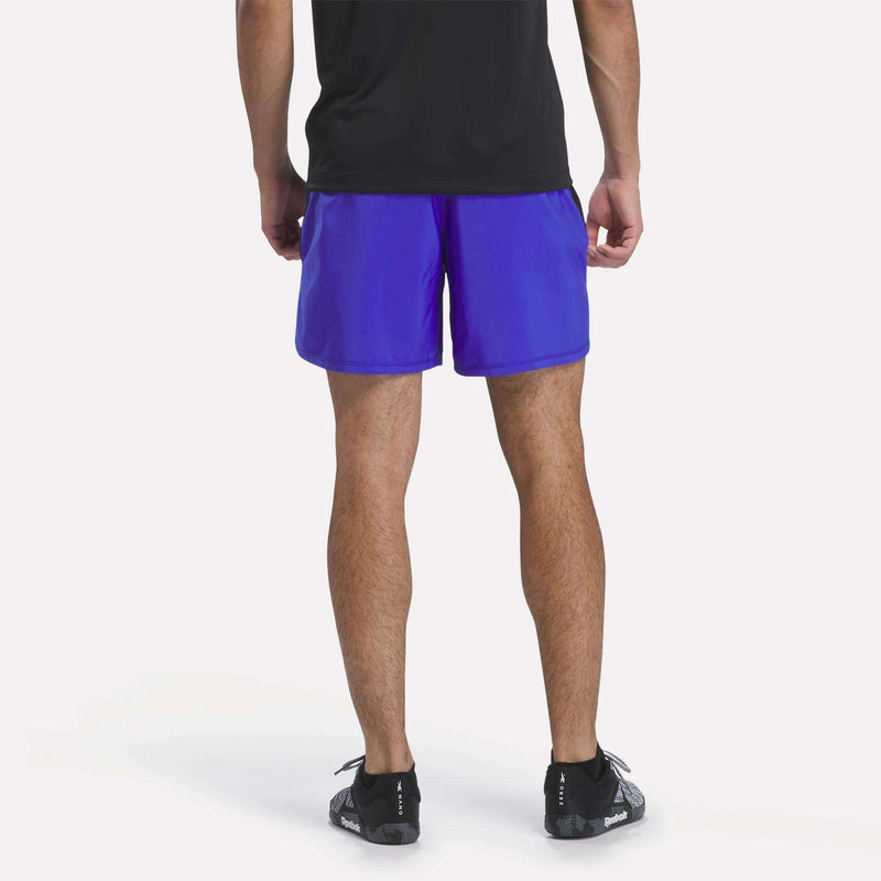A person is shown from the waist down, facing away, wearing a black shirt and Reebok Men’s Running Shorts 7″ in bright blue, paired with black athletic shoes, against a plain light background.