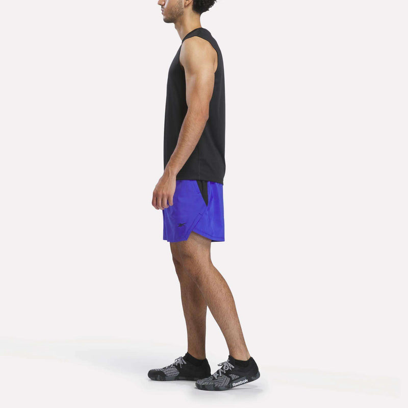 A man in a black sleeveless shirt, black running shoes, and Reebok Men’s Running Shorts 7″ in bright blue stands in profile against a plain white background.